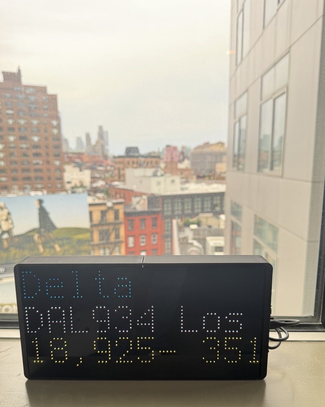 NearestPlane display on a New York windowsill above the rooftops, showing a Delta flight to Los Angeles climbing through 18,925 feet