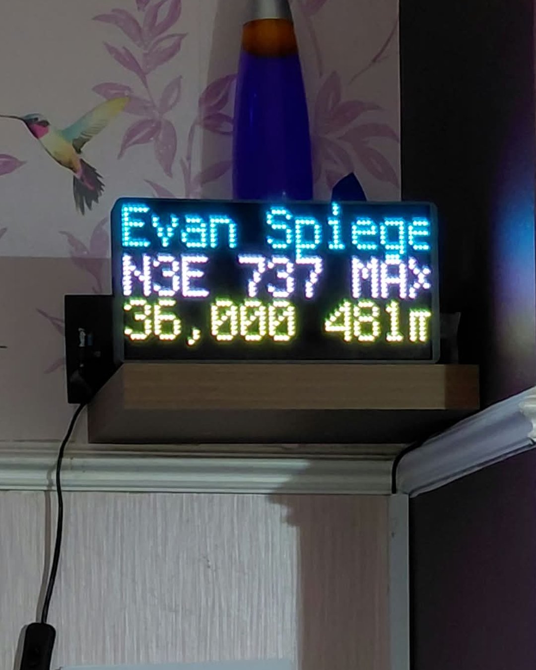 NearestPlane display on a shelf beneath a lava lamp, naming a private jet passing overhead at 36,000 feet